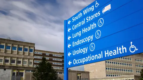 Getty Images Stock photo shows a generic hospital signpost pointing to where the various wards are, with a hospital building in the background against a cloudy sky.