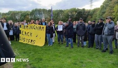 Men and women of different ethnicities stand in a green field, or large area of grass, looking in the same direction as someone, not captured in the image, speaks. Some are holding a large, yellow banner that says Birmingham Black Sisters. Most people have jeans, coats and jumpers on.