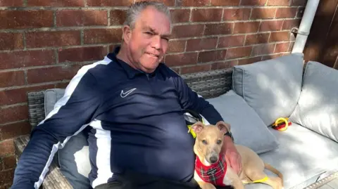 Handout Paul Lockhart sits on a grey outdoor sofa and smiles at the camera, his beige puppy wears a tartan harness and cuddles up to Paul who is wearing a navy and white Nike shell suit top. There is a brick wall behind them.