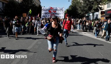 A woman with a megaphone runs towards the camera as the crowd approaches behind her