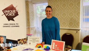 Krystal Keeley, wearing a blue long sleeved top, standing behind a table with interactive toys - a rainbow and stress balls - on. There is a Unique Voice banner to the left of the picture with a slogan, 'using drama to enhance learning'. Behind Ms Keeley there is a wall with cashew style wallpaper and two long windows with a light net curtain over.