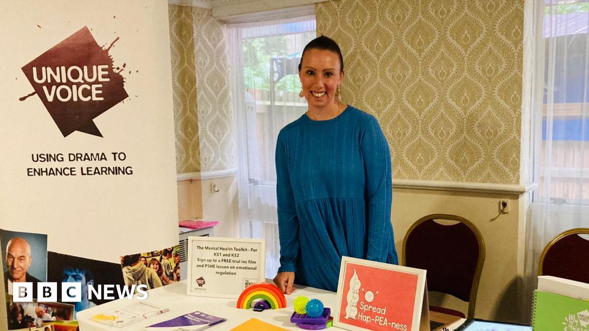 Krystal Keeley, wearing a blue long sleeved top, standing behind a table with interactive toys - a rainbow and stress balls - on. There is a Unique Voice banner to the left of the picture with a slogan, 'using drama to enhance learning'. Behind Ms Keeley there is a wall with cashew style wallpaper and two long windows with a light net curtain over.