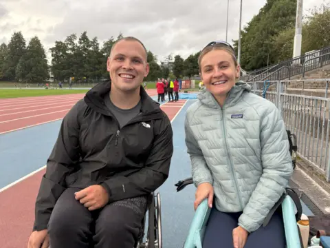 Image of wheelchair racers Ben Rowlings and Melanie Wood at their running track. The pair are smiling and wearing outdoor jackets