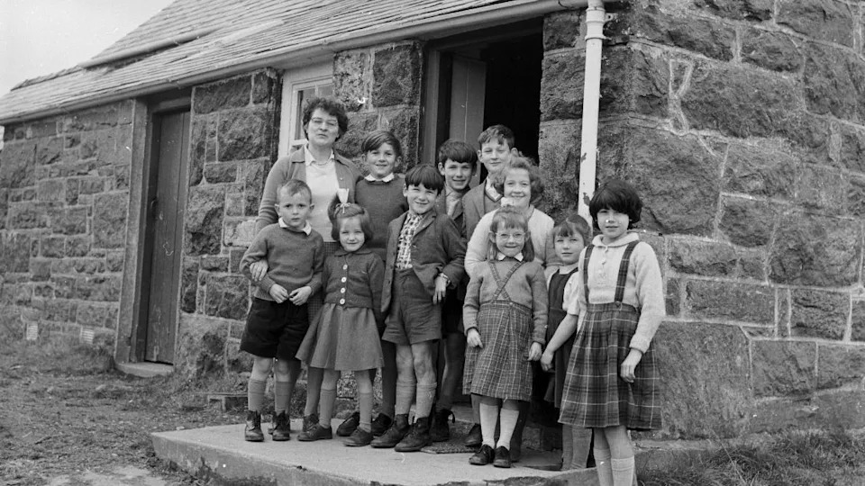 Black and white photograph showing very young school pupils outside the school of Capel Celyn. Ten young school pupils can be seen huddled on the school steps. The boys were shorts, jumpers and shirts. While the girls were tartan skirts and cardigans. Their teacher stands tall at the back of the group, they all smile at the camera. 