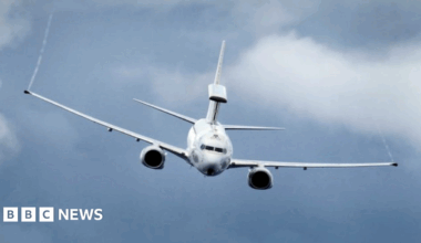 A large, white military aircraft flies in the sky. It has long wings and shorter tail wings and aircraft paraphernalia on top of its rear end.
