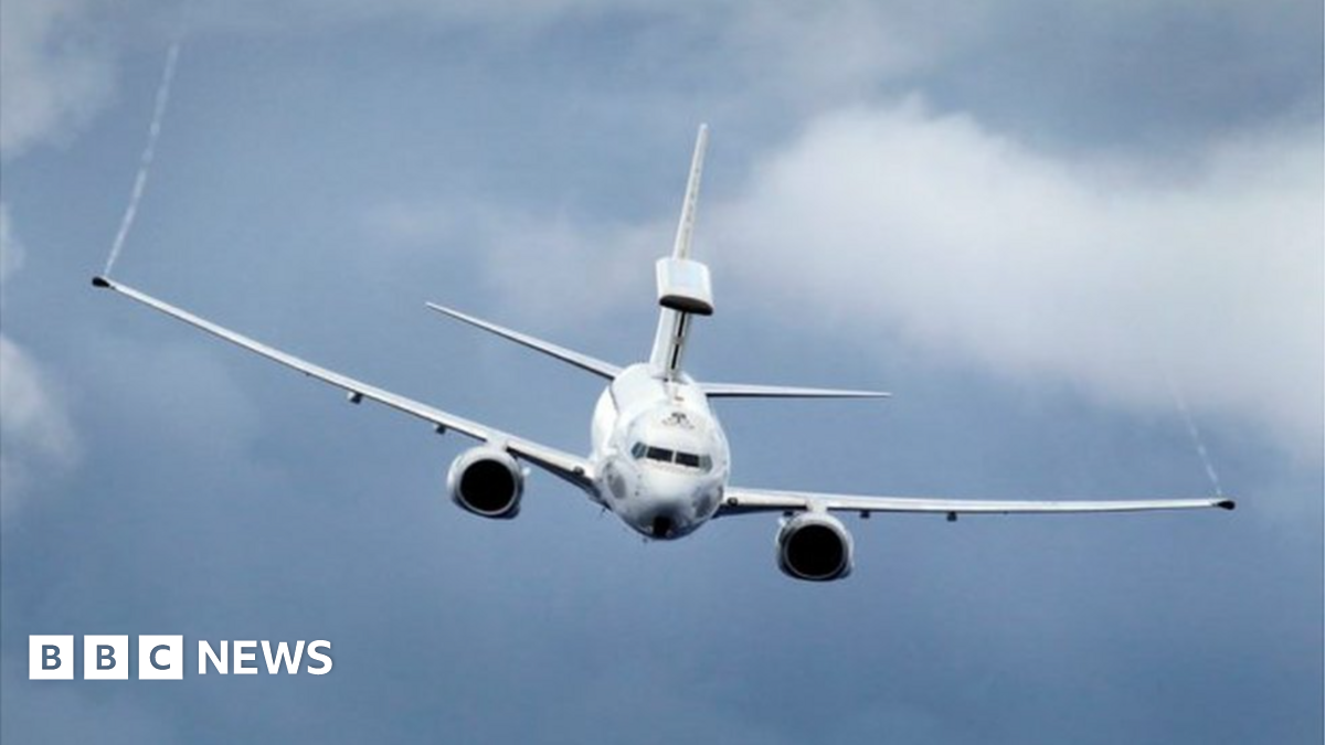 A large, white military aircraft flies in the sky. It has long wings and shorter tail wings and aircraft paraphernalia on top of its rear end.