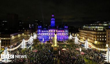 A view from high up, looking down on a city square. Christmas decorations are all around it, along with crowds of people, while the city chambers building behind the square is lit up in purple.