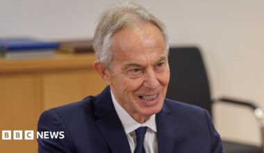 Tony Blair wears a blue suit and tie with a white shirt. He has grey hair and there is a wooden desk behind him.