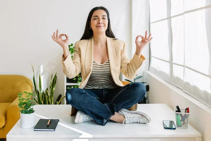 Person sitting cross-legged on a desk in an office, meditating with a relaxed expression