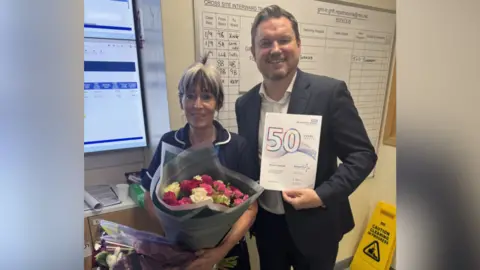 NHS Gloucestershire Hospitals Foundation Trust The Image shows two people; one woman holing two bunches of flowers and a man holding a certificate with a big 50 in the middle. He is wearing a suit and the woman is in a nurses uniform. A cleaning can be seen in the background. 