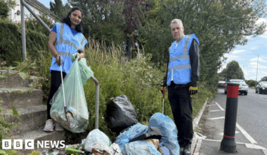 Two people in blue high-vis jackets hold litter-pickers and stand next to piles of blue plastic bin bags. Some have broken open and some items, such as a coffee tin and balsamic vinegar bottle, litter the street.