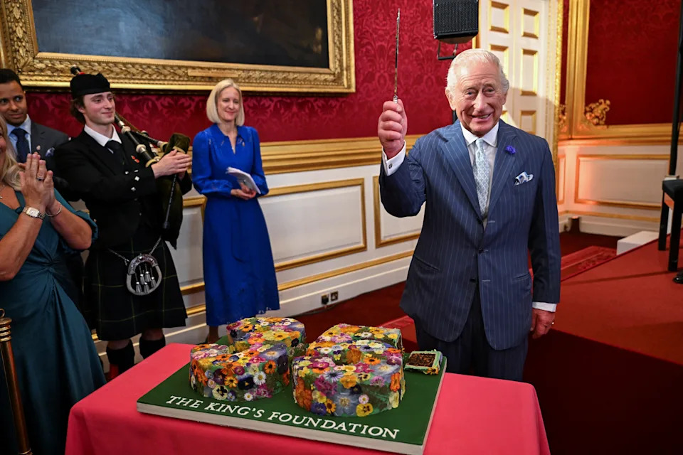 King Charles wearing a blue suit holding a cake knife in front of a cake reading The King's Foundation