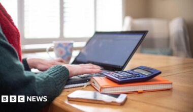 There is a laptop, calculator, mobile phone and notepad on a desk and a woman's arms and hands are typing
