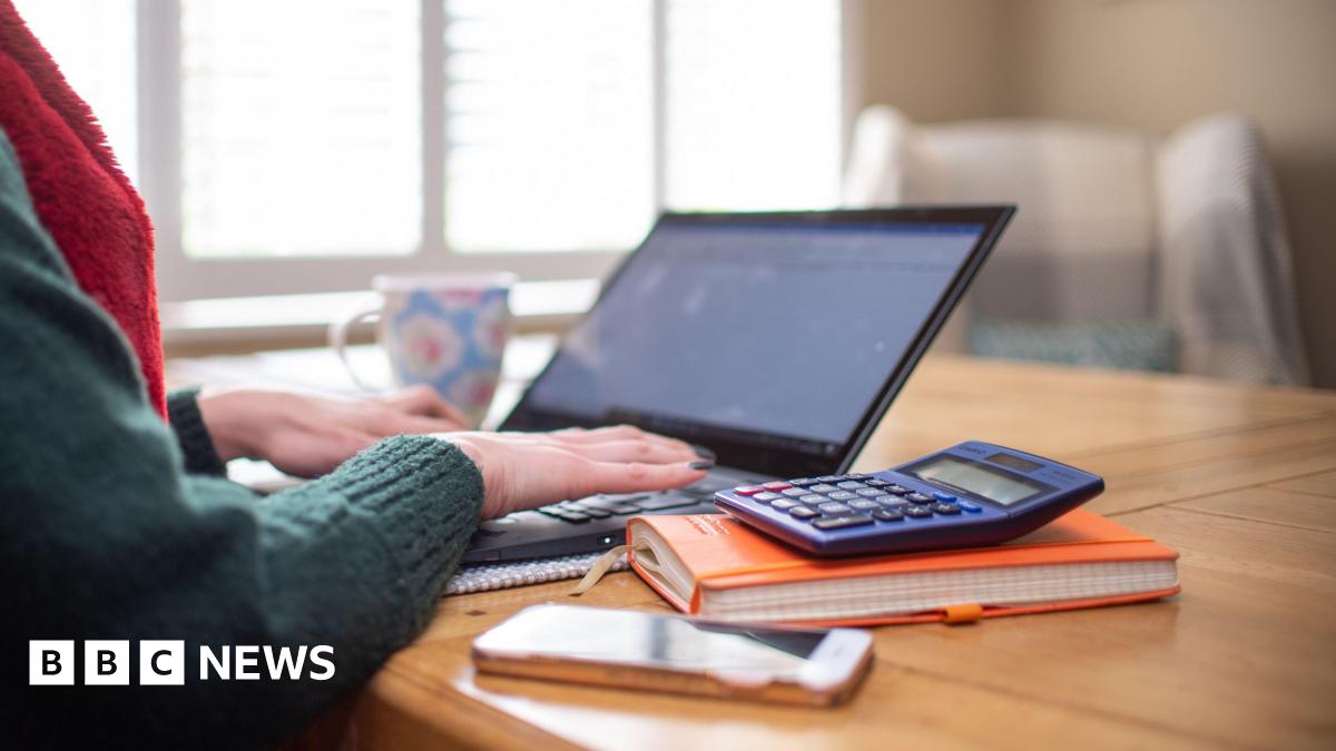 There is a laptop, calculator, mobile phone and notepad on a desk and a woman's arms and hands are typing