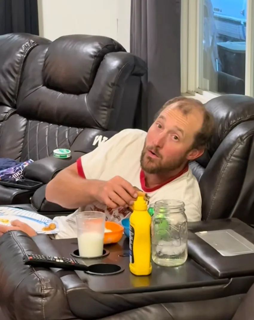 Person relaxing on a recliner, wearing a casual t-shirt, eating snacks, with a drink, mustard bottle, and remote control on the side table