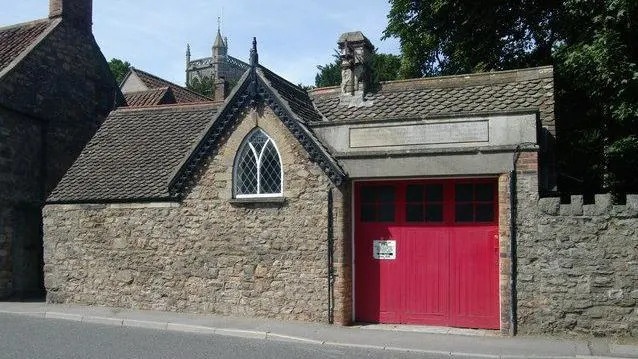 A single story stone cottage with a red glass-panelled door, out of which Emily's horse-drawn fire engine would have exited.