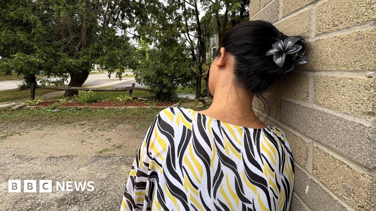 A photo of the woman interviewed in the story. It does not show her face and is taken from behind. Her hair is black and is held up by a black clip shaped like a flower. She is wearing a blouse with yellow, black and white print. She leans on a brick wall, and is overlooking a suburban road lined by trees.