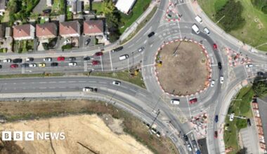 An aerial view of a roundabout on a busy road junction in Pudsey. The road has red and white barriers surrounding it, indicating improvement work is taking place.