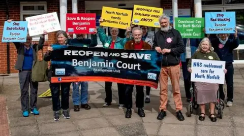 Save Benjamin Court A brick building with a glass entrance with a group of protesters gathered outside holding banners reading "Save Benjamin Court".