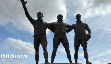 A statue of three men in rugby jerseys with one holding a rugby ball. The statue on the left of the image has one arm in the air while the others lock arms stood side by side. To the right of the image three people can be seen dressed in formal attire.