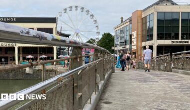 Pero's Bridge in Bristol. There are railways either side of the pathway. There are some padlocks hanging on it. In the distance, people can be seen walking around and there are restaurants and a white ferris wheel.