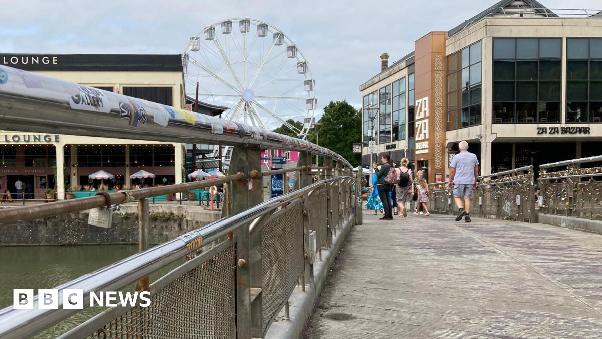 Pero's Bridge in Bristol. There are railways either side of the pathway. There are some padlocks hanging on it. In the distance, people can be seen walking around and there are restaurants and a white ferris wheel.