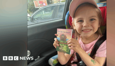 A young girl sits in a car seat smiling at the camera as she holds a book. She is wearing a pink cap and a pink T-shirt. A green fake tattoo is on her forearm in the shape of an octopus.