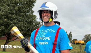A man in a blue t-shirt with a white helmet on. He is carrying a gold baton. He is smiling and the words Baton of Hope Tour 2025 are on the front of his shirt