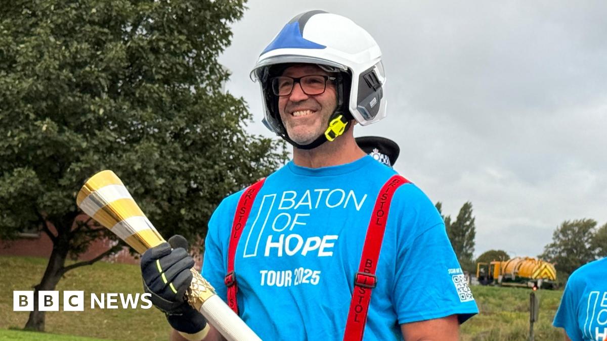 A man in a blue t-shirt with a white helmet on. He is carrying a gold baton. He is smiling and the words Baton of Hope Tour 2025 are on the front of his shirt
