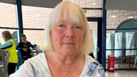 BBC Judith Marshall, a middle-aged woman with bobbed blonde hair looks at the camera. She is wearing a white top with flowers in shades of blue and a gold  and jade necklace. Behind her is the reception area of Scotstoun pool, a light area with full-length windows, some tables and chairs and a fire extinguisher sits to the side.