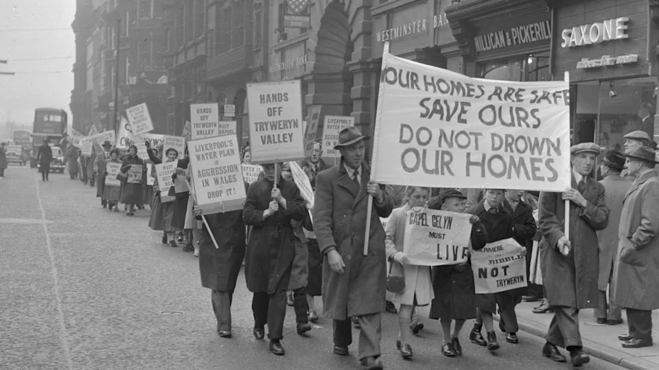 Black and white photograph of protesters walking the streets in Liverpool. Men, women, and children can be seen holding a large poster at the front of a line of protesters which reads: 'Your homes are safe. Save ours. Do not drown our homes'. Other posters read: Hands off tryweryn valley. Liverpool's water plan is aggression in Wales. 