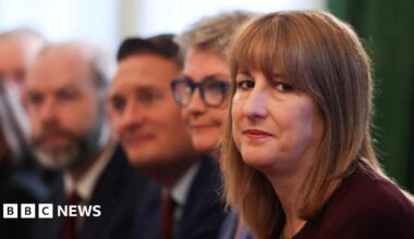Rachel sits on the right of the frame, wearing a dark maroon top, looking seriously into the camera. Behind her sit several cabinet ministers, who appear blurry.