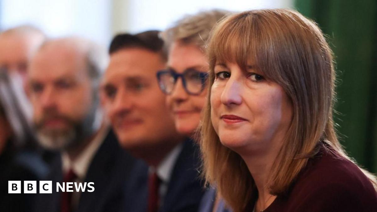 Rachel sits on the right of the frame, wearing a dark maroon top, looking seriously into the camera. Behind her sit several cabinet ministers, who appear blurry.