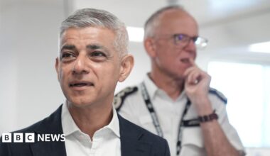 The mayor of London is wearing a dark navy suit, white shirt, no tie and is standing in front of the Met Police Commissioner, Sir Mark Rowley - he is wearing a white police shirt with epaulettes on the shoulders and a lanyard around his neck.