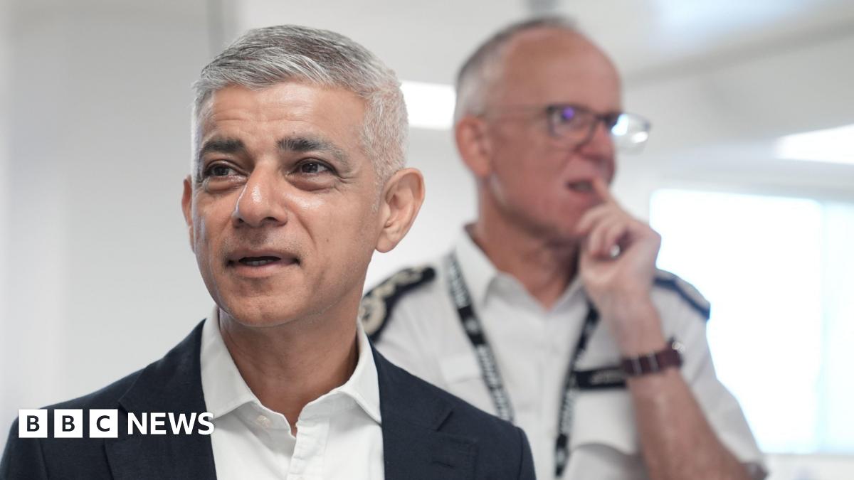 The mayor of London is wearing a dark navy suit, white shirt, no tie and is standing in front of the Met Police Commissioner, Sir Mark Rowley - he is wearing a white police shirt with epaulettes on the shoulders and a lanyard around his neck.