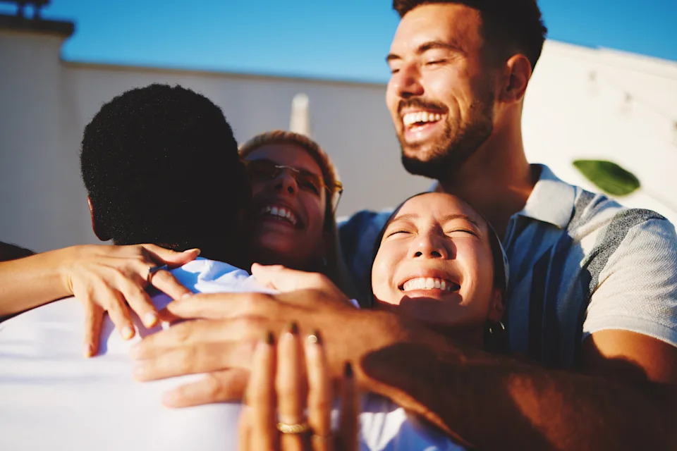 Four people joyfully embrace outdoors, all smiling and basking in the sunlight, conveying happiness and connection
