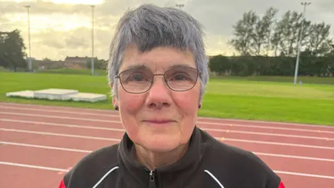 Athletics coach Caroline Johnston - a woman with grey hair and glasses - standing on a running track, with grass behind her and houses beyond that. She is wearing a black tracksuit with white and red trim.