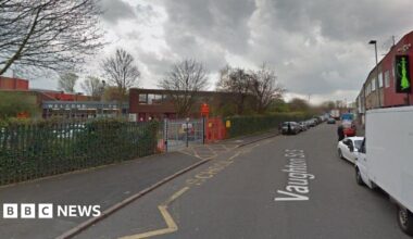 Vaughton Street South in Birmingham. There is a row of parked cars on both sides of the road. In the foreground is a school, with yellow markings on the ground indicating its entrance. There is red signage at the gate.