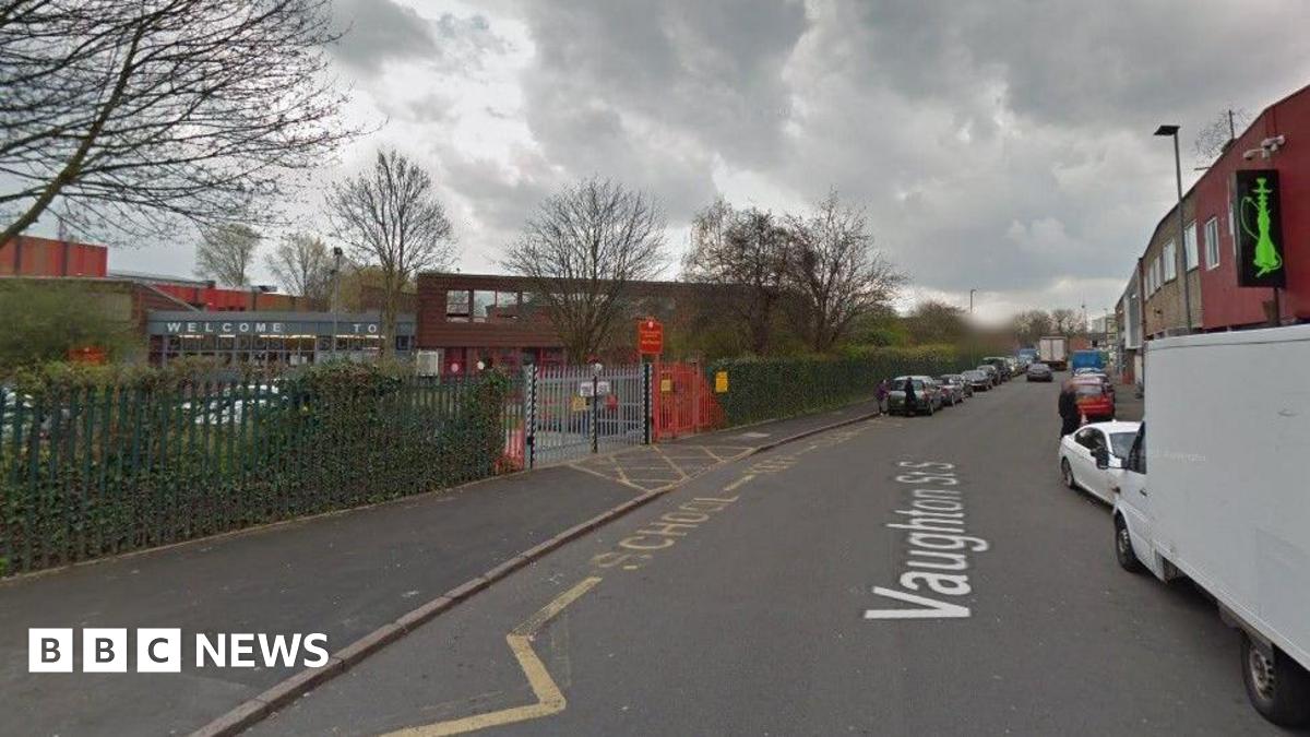 Vaughton Street South in Birmingham. There is a row of parked cars on both sides of the road. In the foreground is a school, with yellow markings on the ground indicating its entrance. There is red signage at the gate.