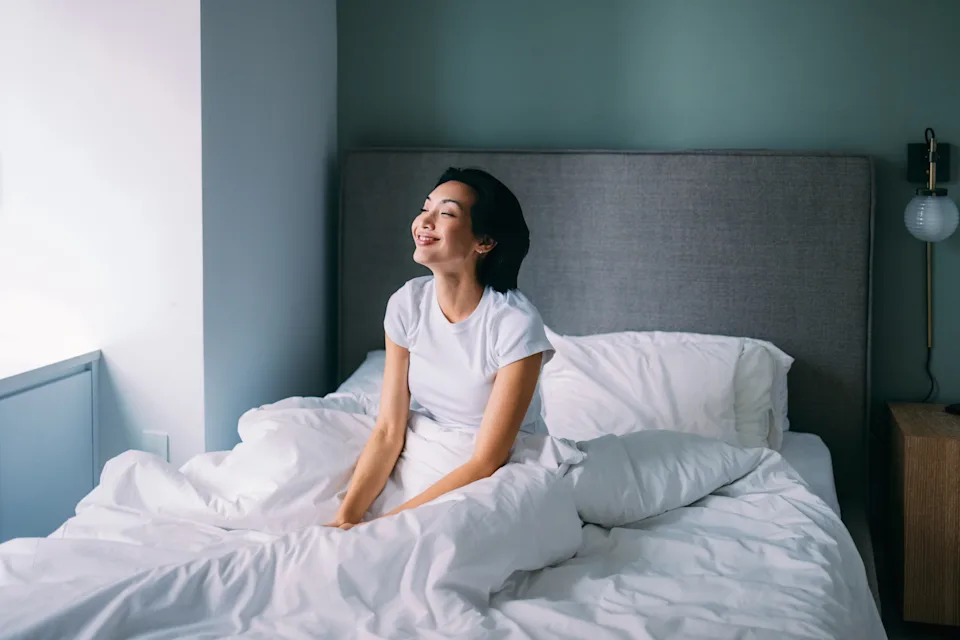 Person in a white t-shirt sitting on a bed, looking out a window, appearing relaxed and content. Room has a minimalist style