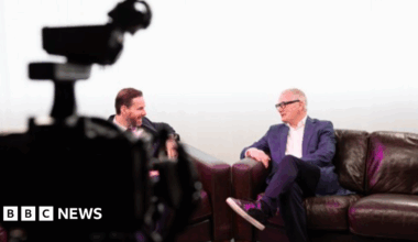 Mayor Richard Parker is sat crossed legged on a brown leather sofa in a television studio that is brightly lit and has white curtains draped in the back. He is sat opposite a man with slicked back brown hair. Mr Parker has grey short hair, wears black framed glasses, a white shirt underneath a blue blazer jacket.