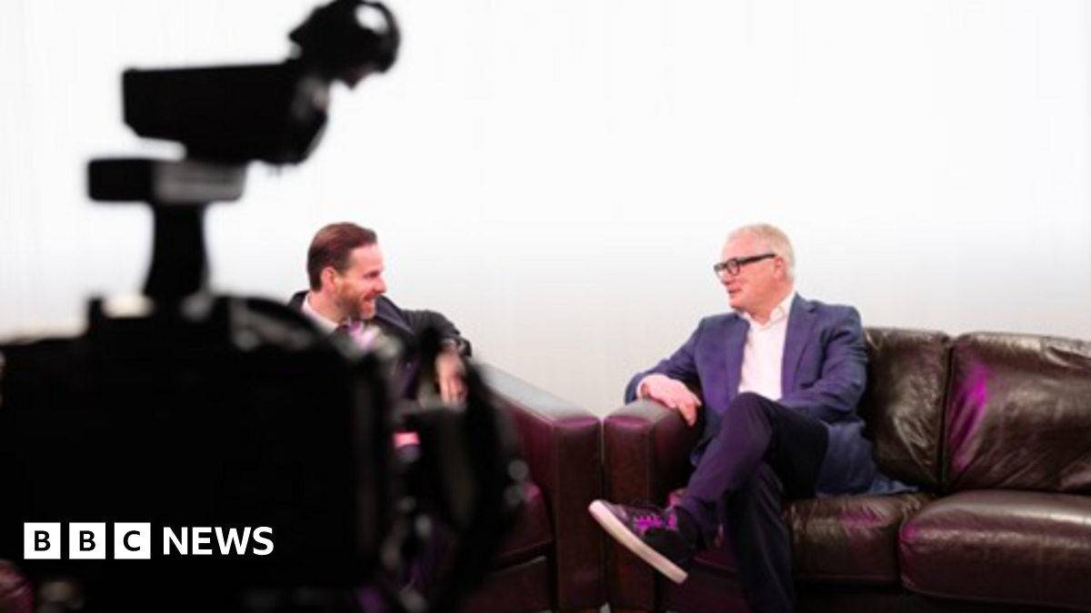 Mayor Richard Parker is sat crossed legged on a brown leather sofa in a television studio that is brightly lit and has white curtains draped in the back. He is sat opposite a man with slicked back brown hair. Mr Parker has grey short hair, wears black framed glasses, a white shirt underneath a blue blazer jacket.