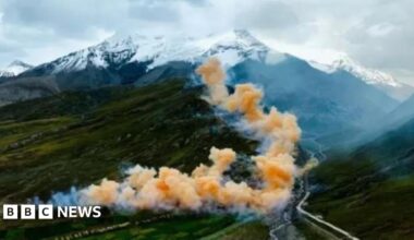 Clouds of colourful smoke rise from a fireworks display over hills, with the snow-capped peaks of the Himalayas in the distance