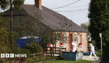 Forensic investigators in white hazmat suits entering a detached stone cottage on a rural road in County Louth.  A blue tent has been erected at the side of the house and there is a blue skip at the front of the property.  The red-framed front door is open.  There are red flower boxs on the front windowsills.