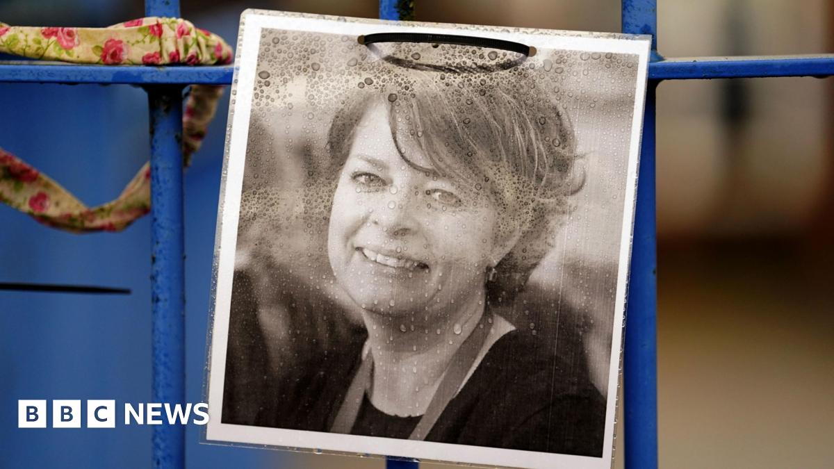 A printed black and white photograph of Ruth Perry fixed to a blue-coloured fence. Mrs Perry is smiling in the photo.