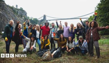 A group of 18 litter pickers - of different ages and genders - stand joyfully with their arms in the air, with the Clifton Suspension Bridge in the background. They are wearing gloves and there are black bin bags on the floor, filled with what appears to be rubbish.