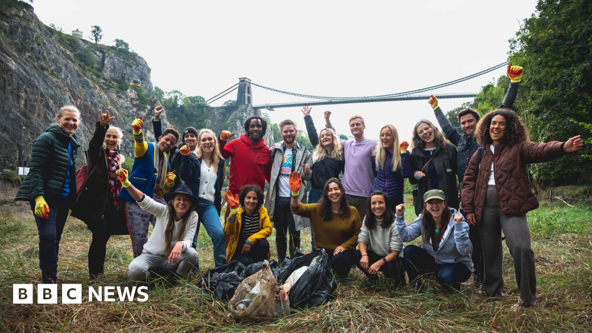 A group of 18 litter pickers - of different ages and genders - stand joyfully with their arms in the air, with the Clifton Suspension Bridge in the background. They are wearing gloves and there are black bin bags on the floor, filled with what appears to be rubbish.