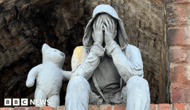 A close up view of a grey sculpture depicting a seated, hooded male figure with hands covering its face, accompanied by a bear-like companion offering comfort with a paw on the figure’s shoulder.
