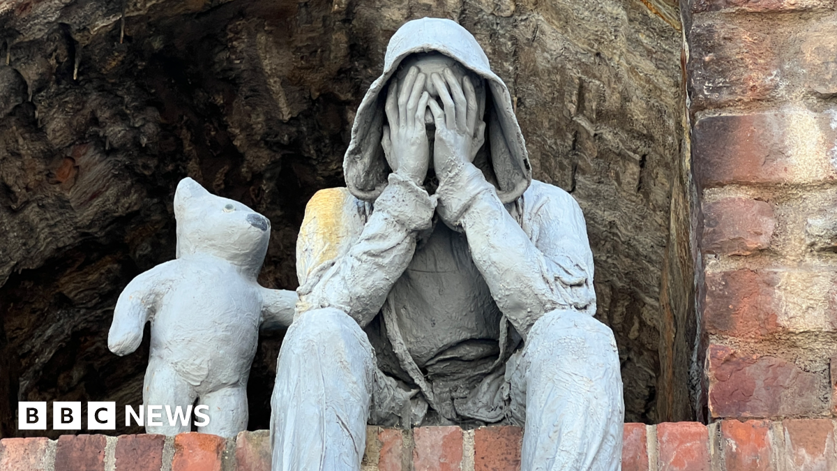A close up view of a grey sculpture depicting a seated, hooded male figure with hands covering its face, accompanied by a bear-like companion offering comfort with a paw on the figure’s shoulder.