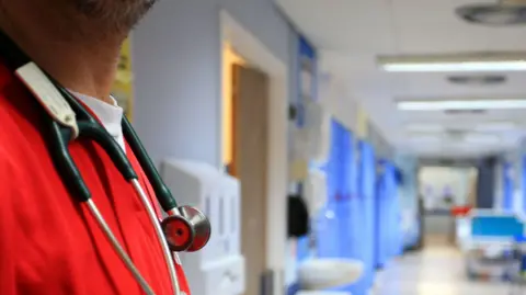 PA A stock image of a man's upper body, cropping out his face, showing him wearing a stethoscope and red scrubs. In the background is a blurred hospital ward.